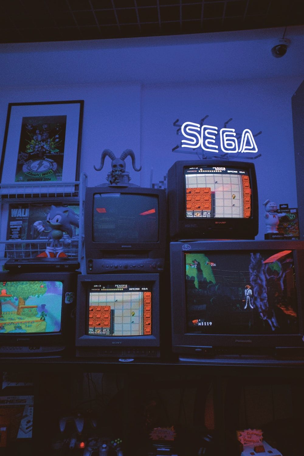 A room with a neon sign that says SEGA and many TVs - Arcade