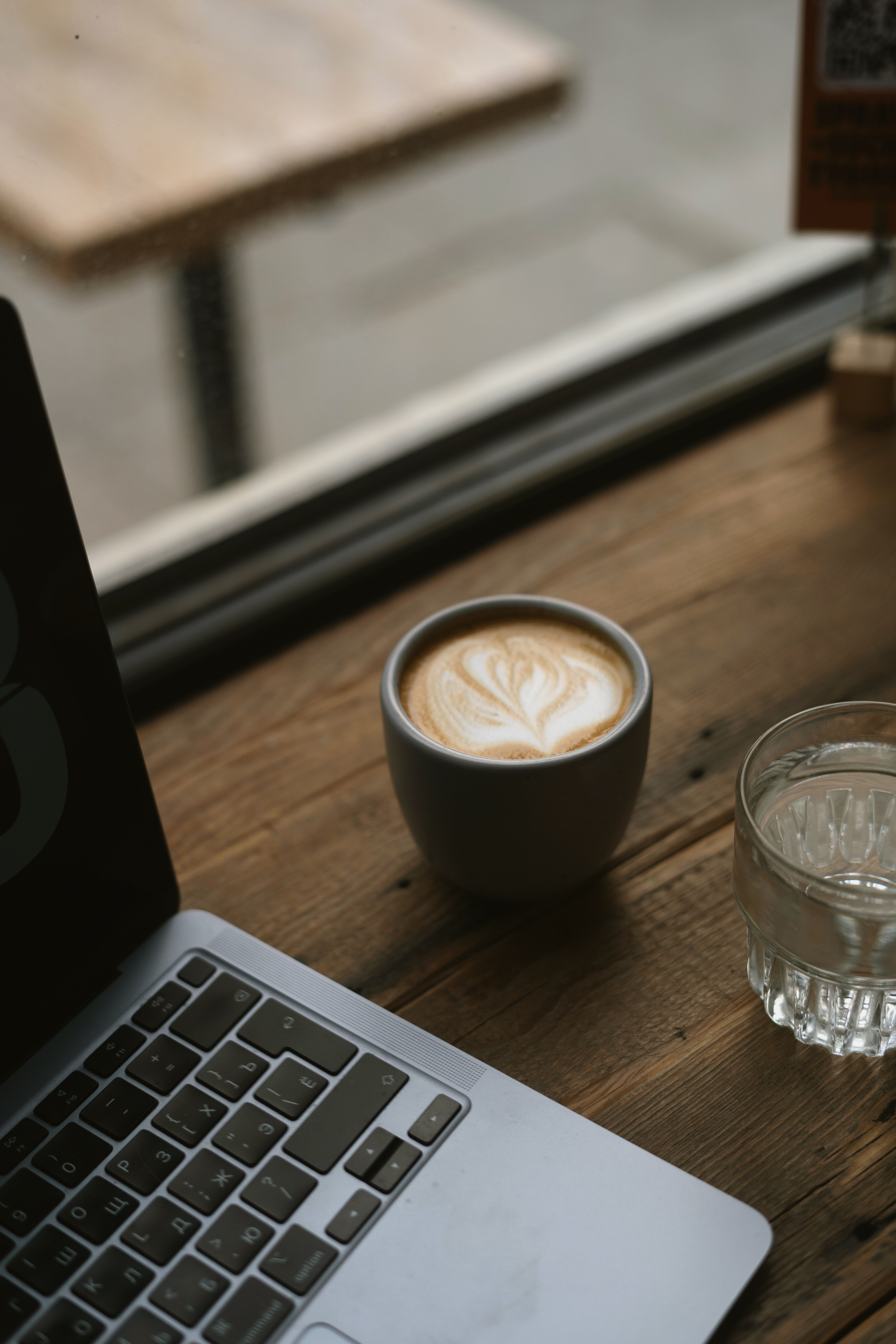 A cup of coffee next to a laptop on a wooden table. - Coffee