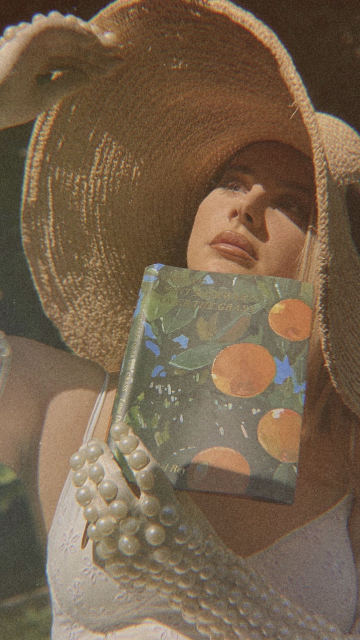 A woman in a sunhat and pearls holds a book with a fruit pattern. - Lana Del Rey