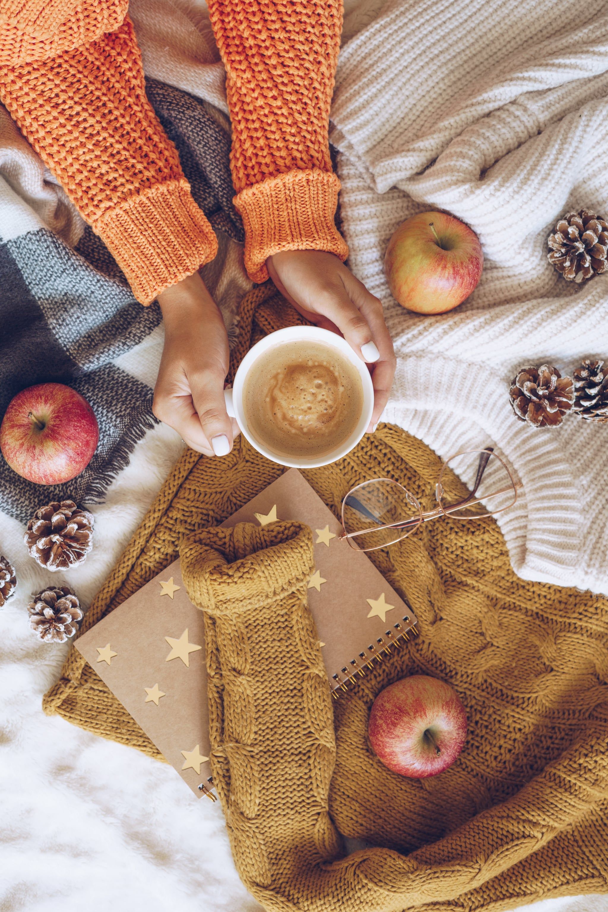 A woman holding a cup of coffee in a cozy sweater surrounded by apples and pinecones. - Cozy