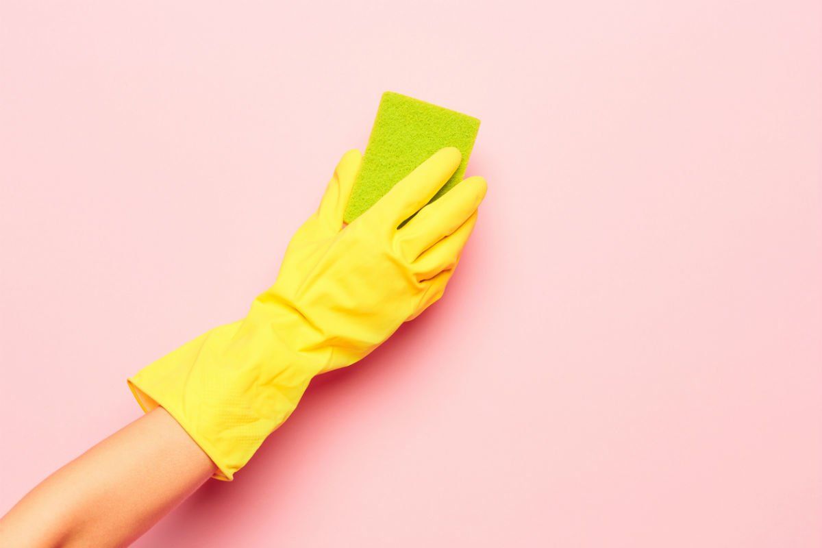 A hand wearing a yellow rubber glove holding a green sponge against a pink background - Clean