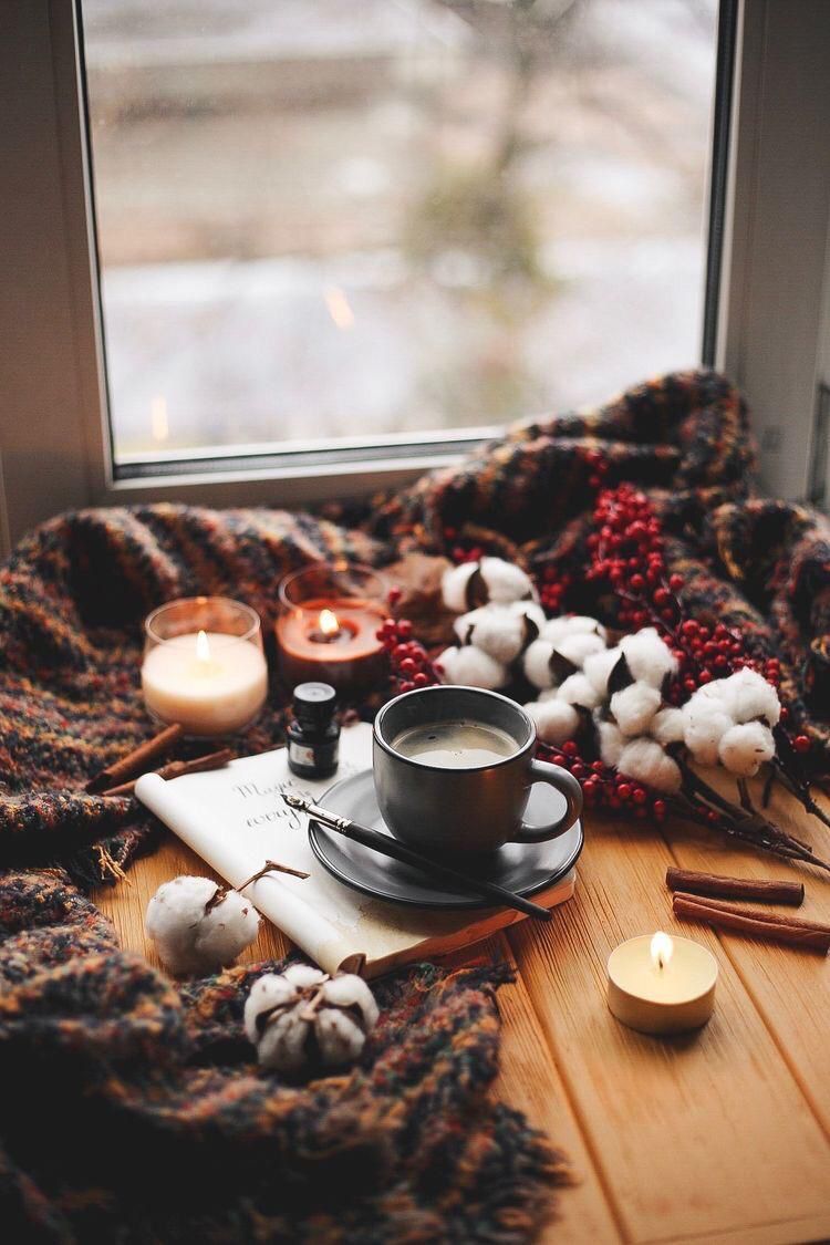 A cup of coffee sits on a table next to a book, surrounded by candles and a blanket. - Cozy