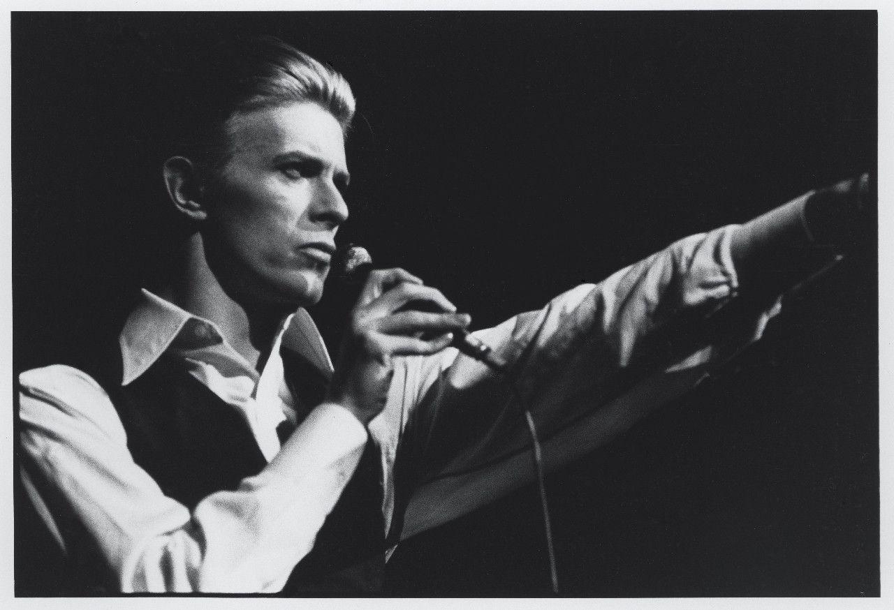 A man holding up his hand while singing - David Bowie