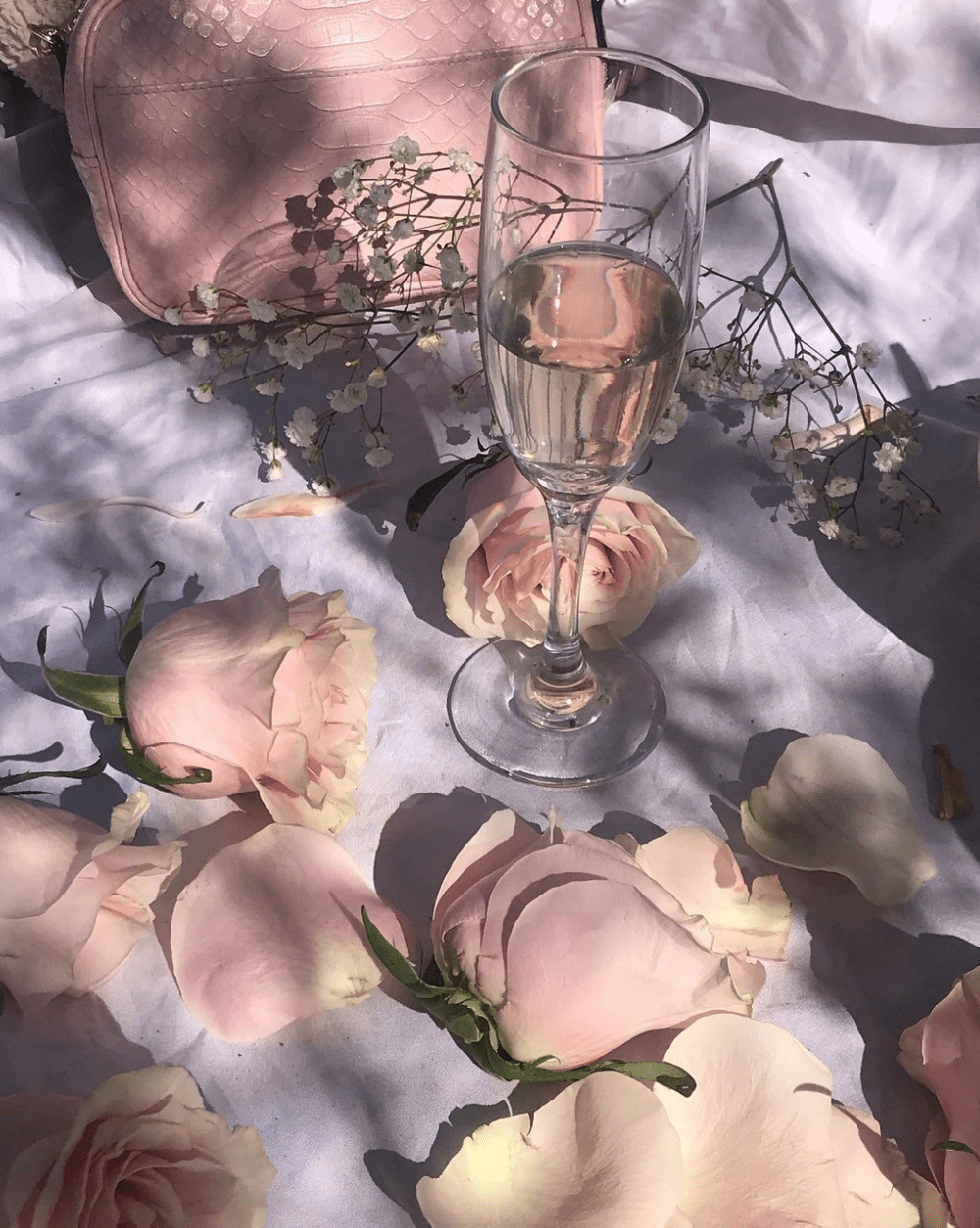 A glass of champagne sits on a table with pink roses and petals. - Champagne