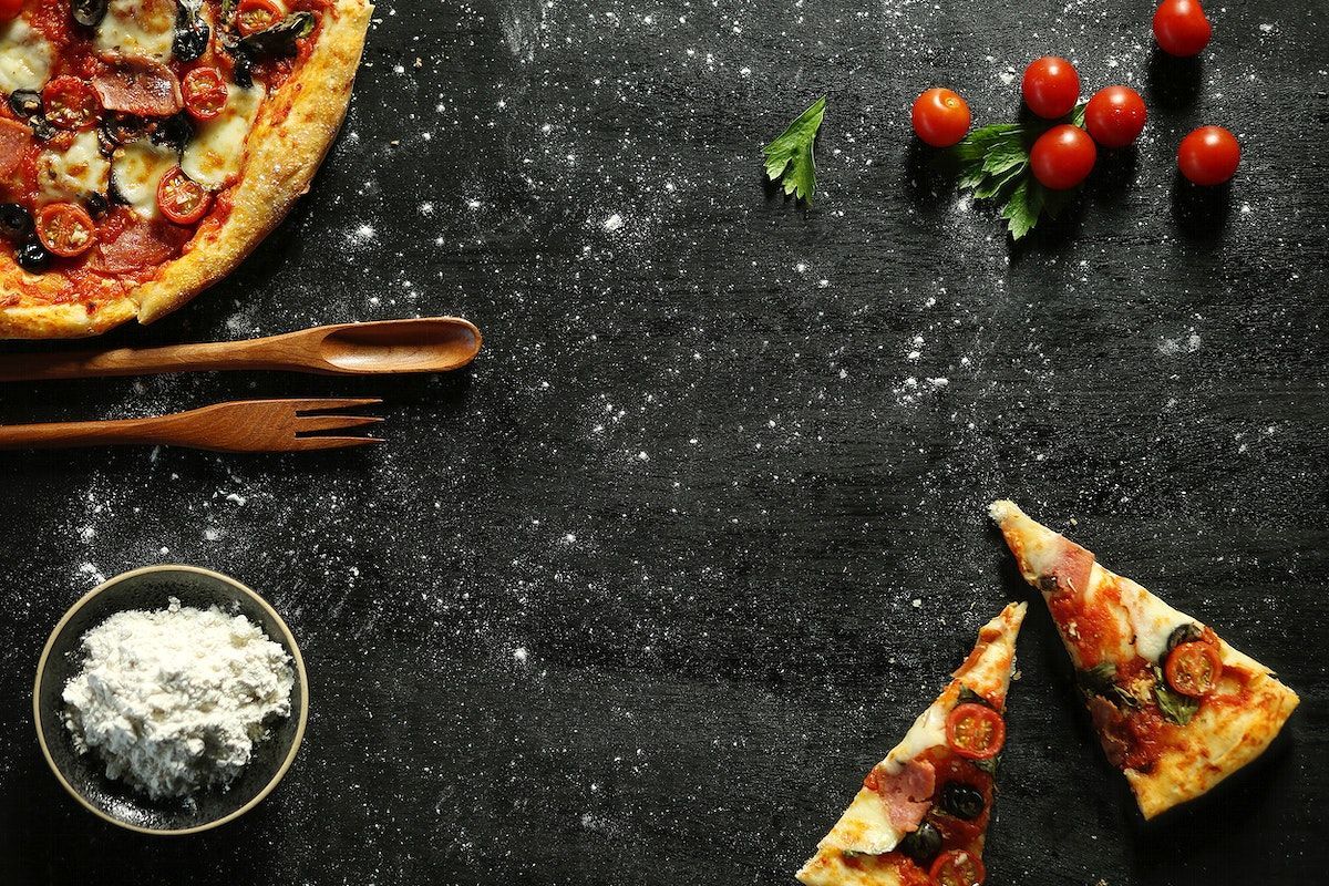 A pizza with a slice cut out, placed on a black surface with a bowl of cheese and tomatoes. - Pizza