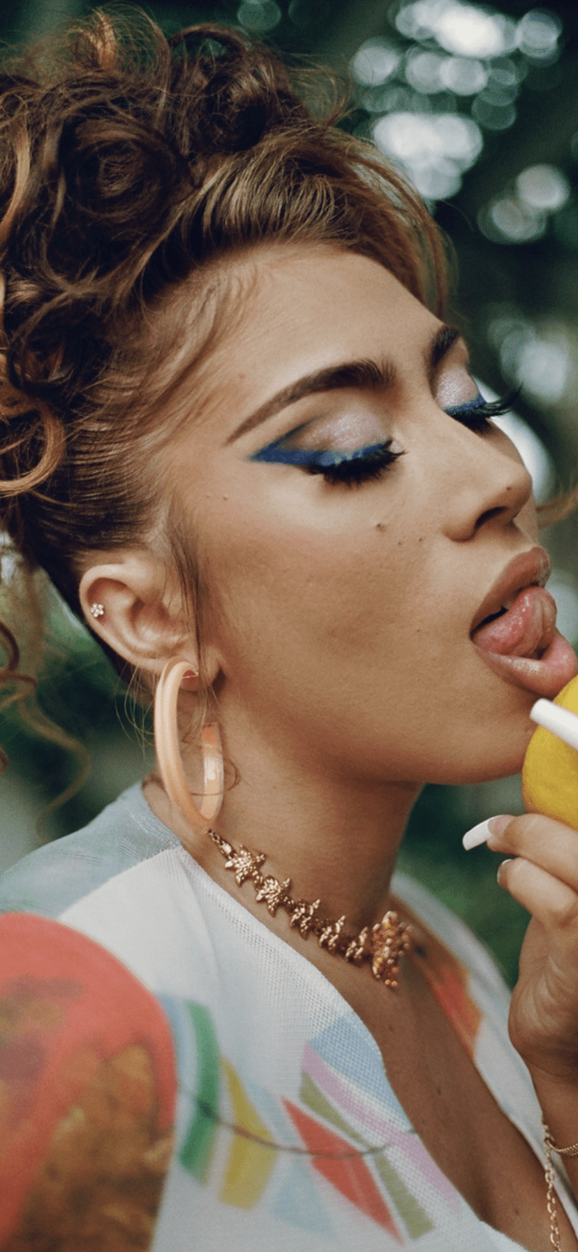 A person with blue eye shadow and gold jewelry eating an apple. - Kali Uchis