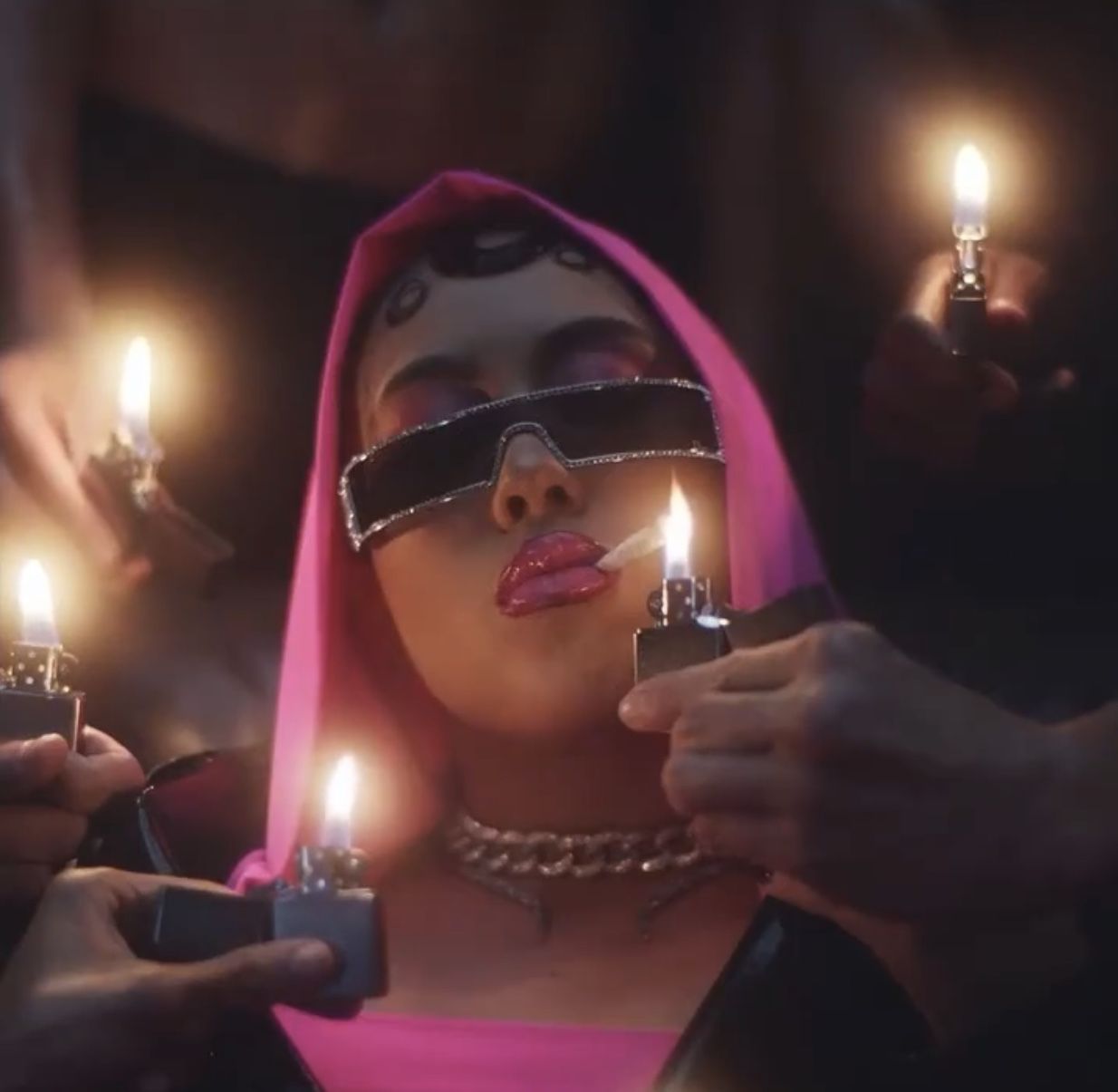 A woman wearing sunglasses and a pink hooded jacket lights a candle - Kali Uchis