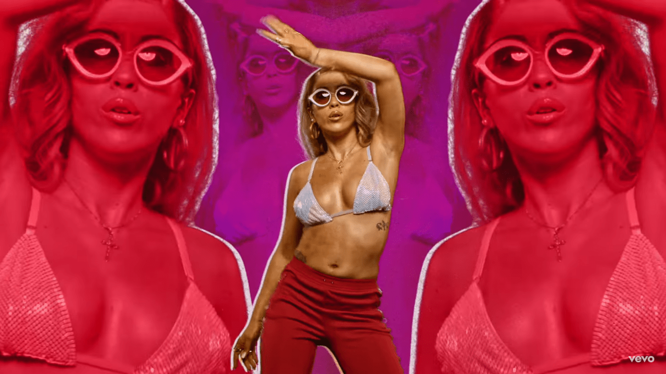 A woman in a bikini top and red pants dances in front of a purple background. - Kali Uchis