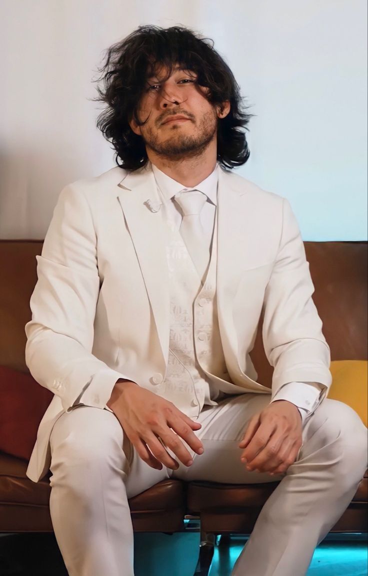 A man with long hair and a white suit sitting on a brown couch. - Markiplier