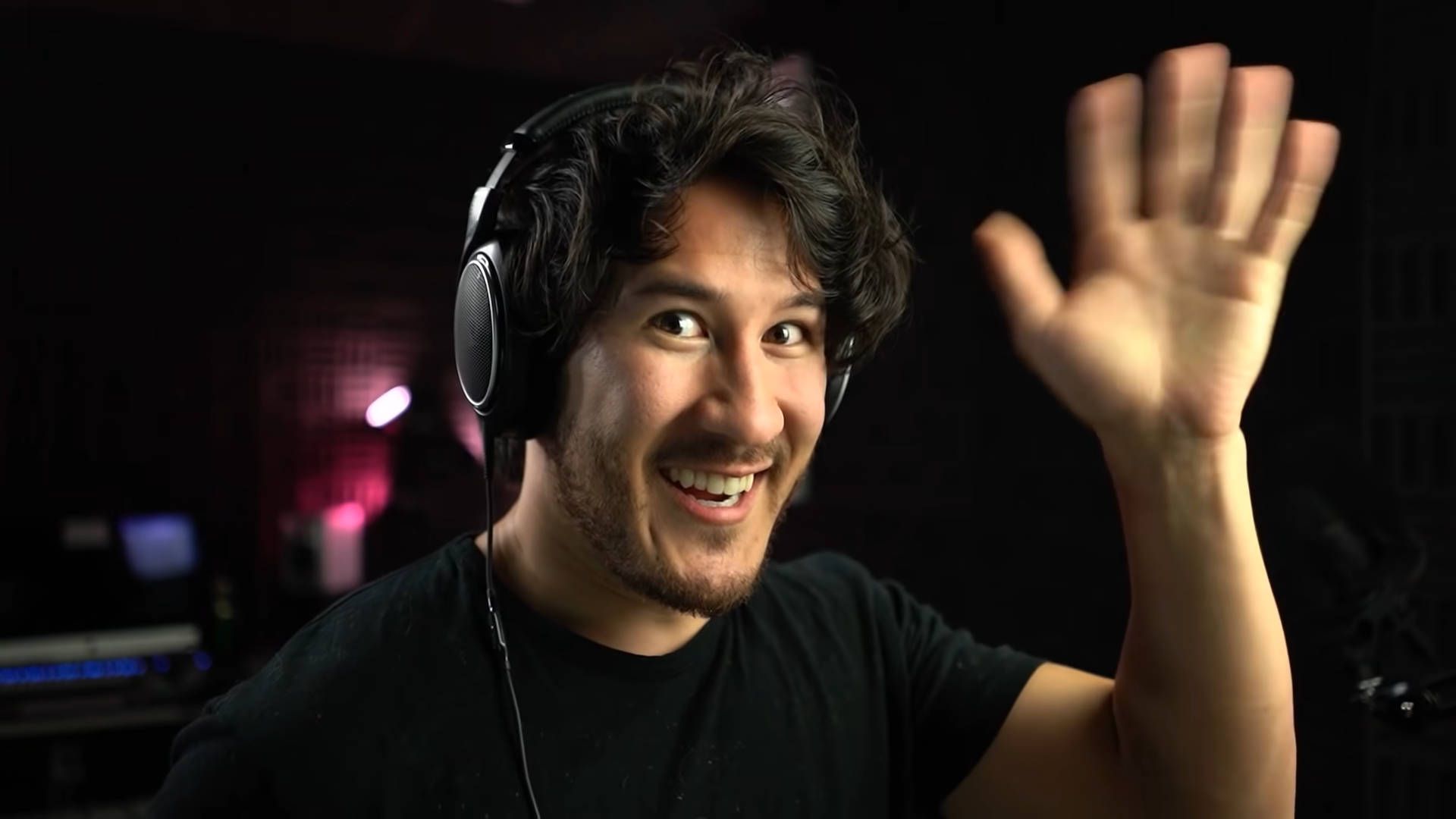 A man wearing a black shirt and a headset smiles and waves at the camera. - Markiplier