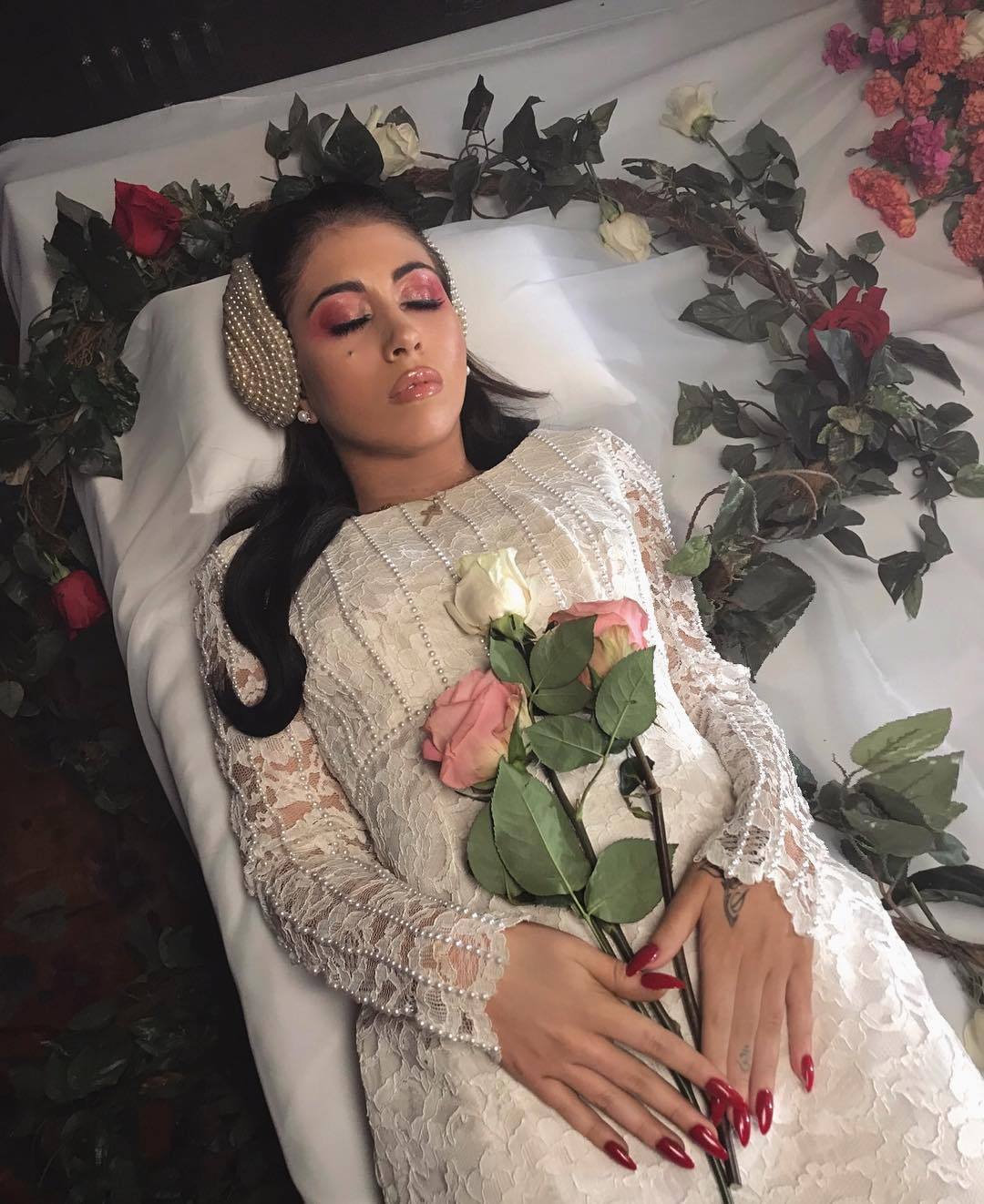 A woman in a white dress lying in a casket holding flowers - Kali Uchis