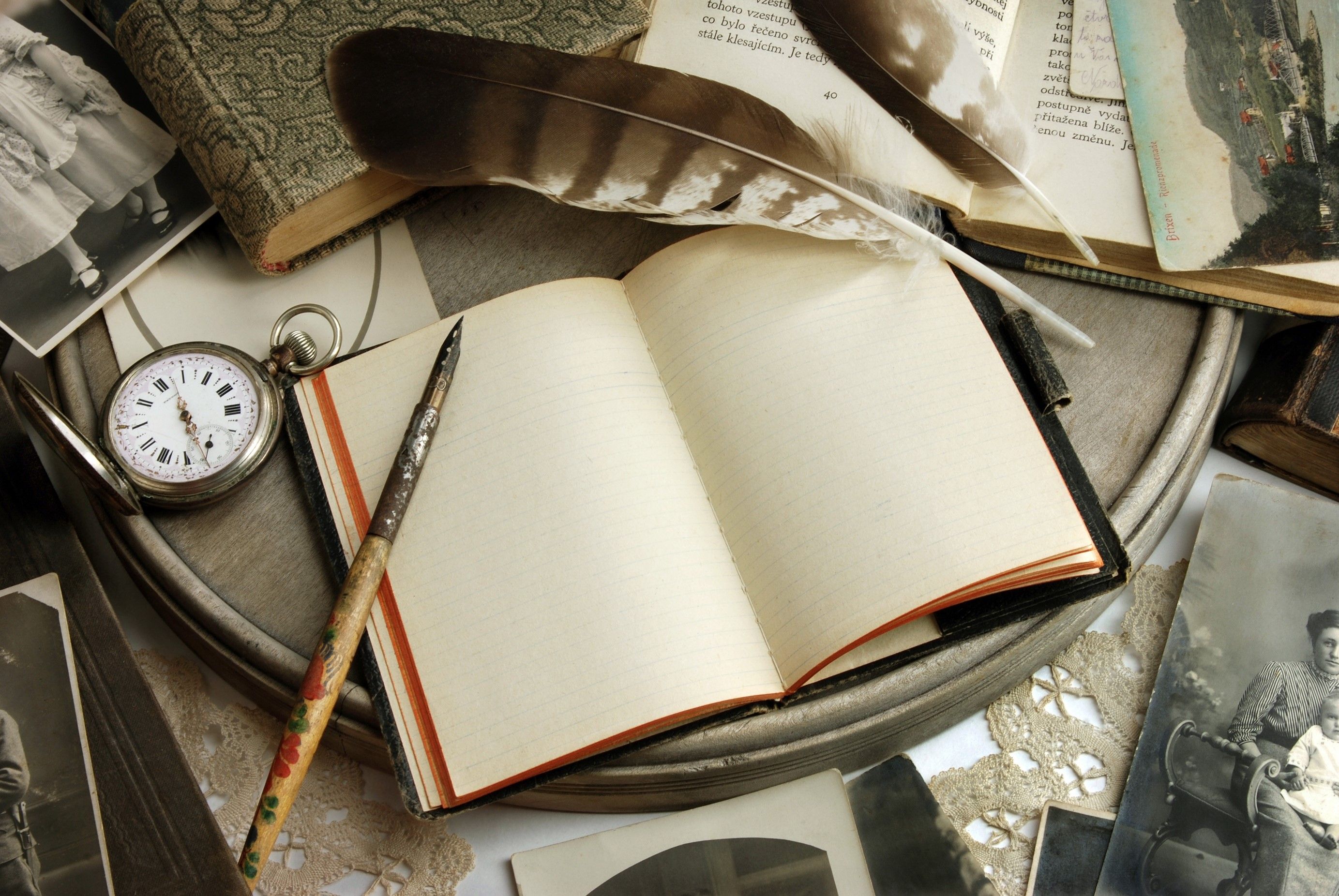 An open notebook, a pen, a pocket watch, and a feather quill sit atop a table with old photographs. - Books