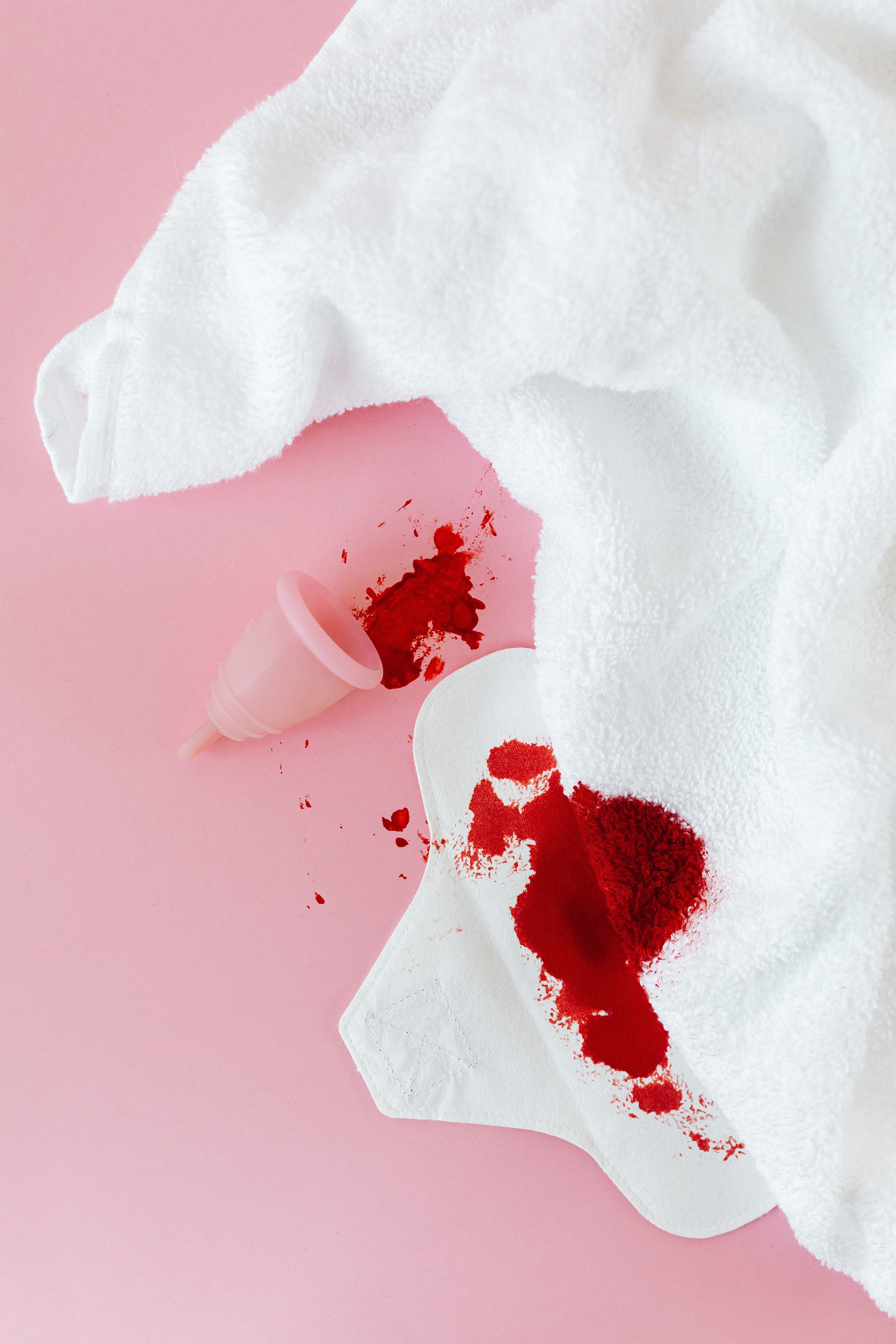 A menstrual cup is seen spilling blood onto a white towel on a pink background. - Blood