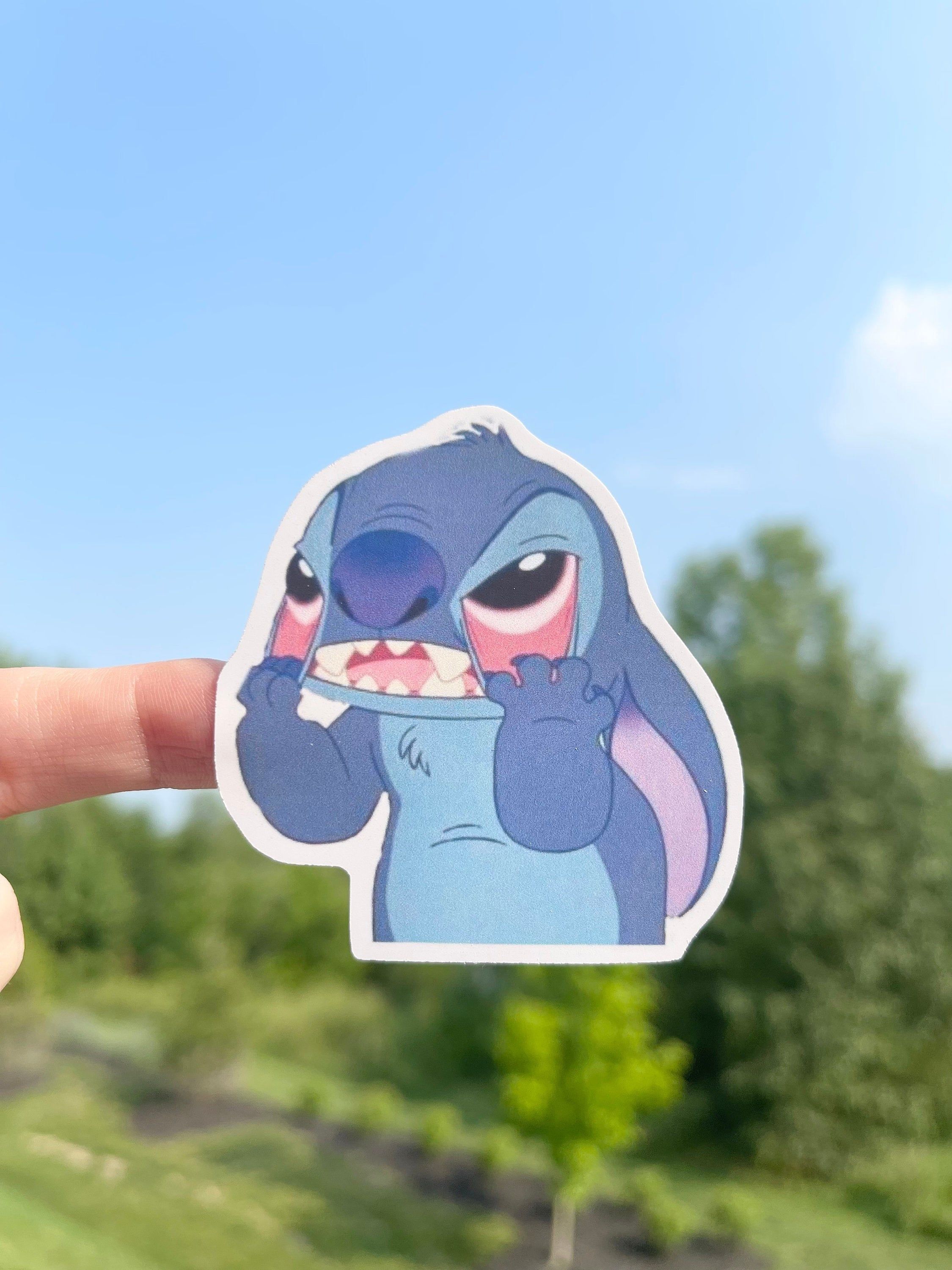 A hand holding a sticker of a cartoon character, Stitch from Lilo and Stitch, holding a shark tooth. - Stitch