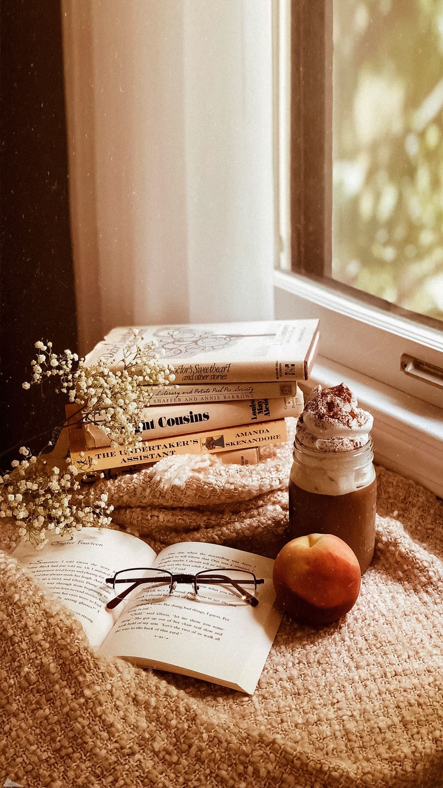 A book, a cup of coffee, a peach, and a pair of glasses on a blanket in front of a window. - Cozy