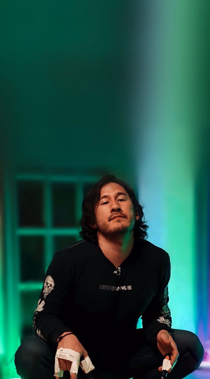 A man with long hair and a beard sitting on the ground with a green light behind him - Markiplier