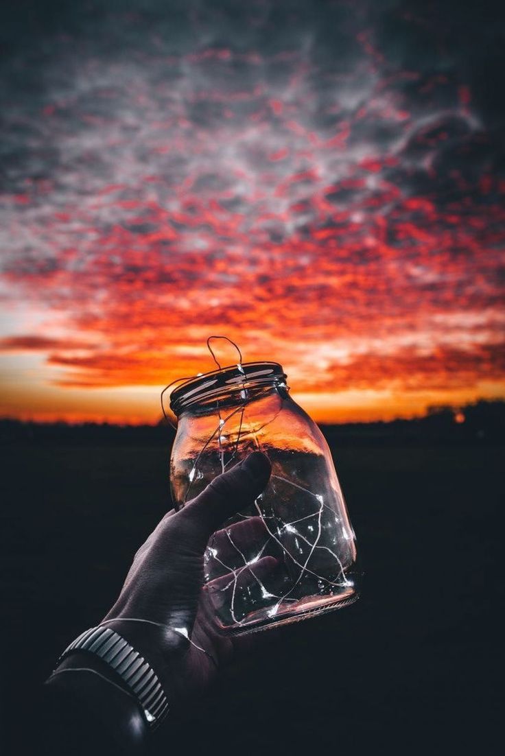 A hand holding a jar with lights inside in front of a sunset. - Photography