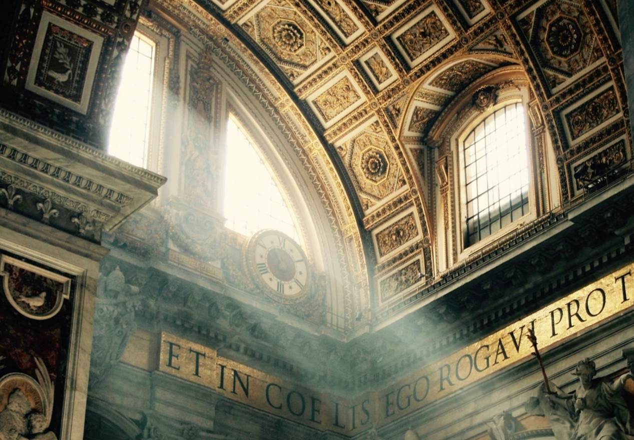 A photograph of the ceiling of St. Peter's Basilica in Rome, Italy. The ceiling is painted gold and features many intricate details. The photograph is taken from the ground looking up, and there is a beam of light shining down through a window. - Dark academia, light academia, architecture