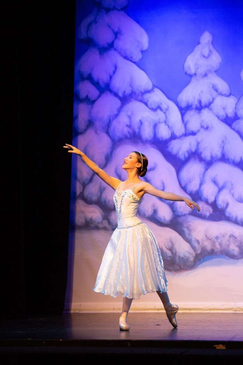 A ballerina in a white dress and white tights stands on stage in front of a snowy background. - Ballet