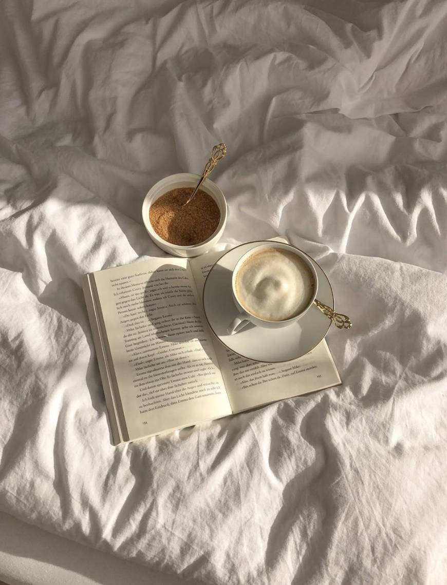 A cup of coffee and a bowl of sugar on top of an open book on a bed. - Coffee