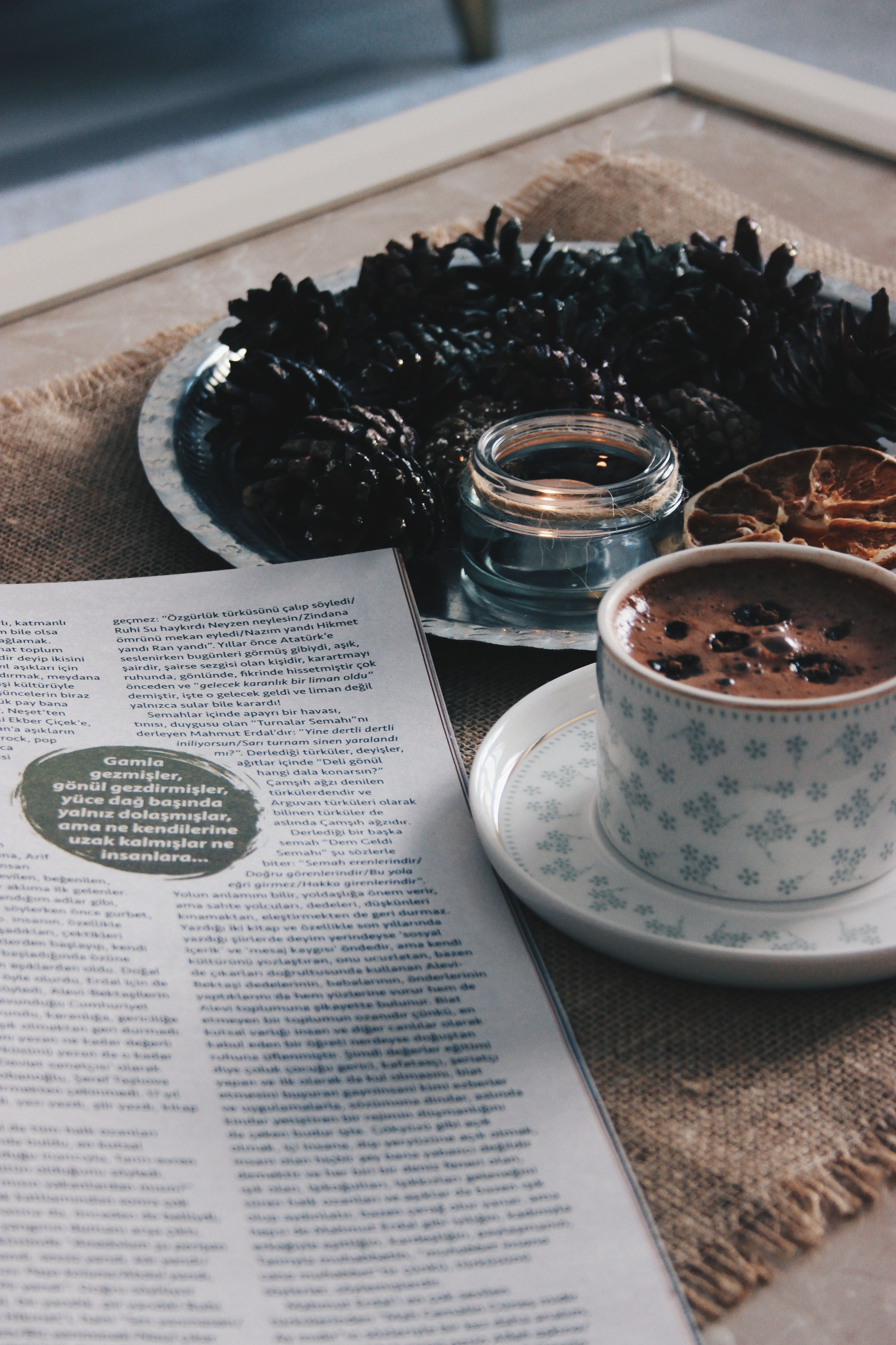 A cup of coffee with a saucer, a plate of coffee beans, a candle, and an open magazine on a table. - Coffee