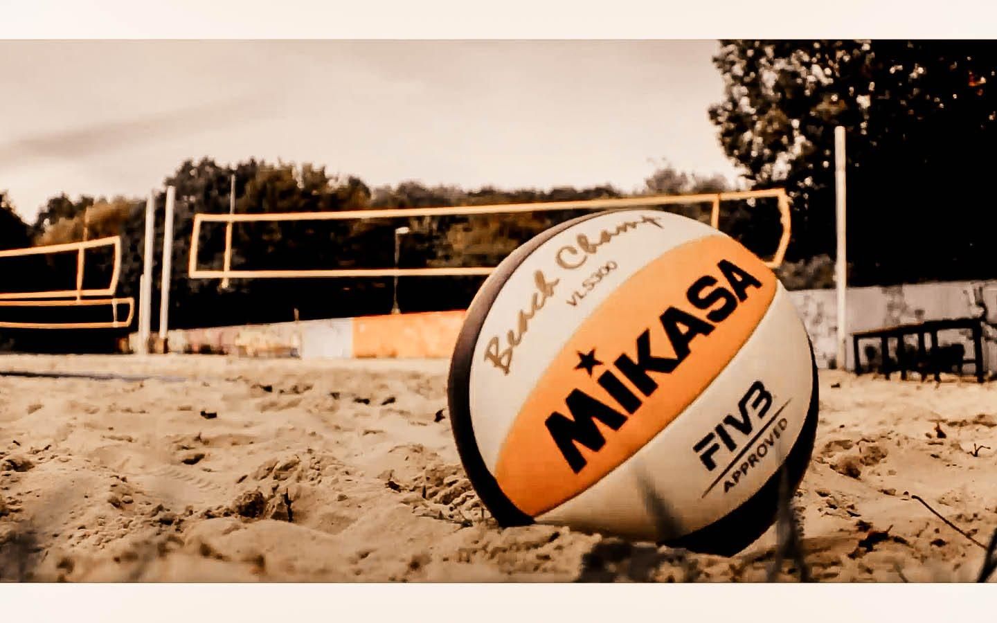 A volleyball sitting on the sand next to an outdoor net - Volleyball