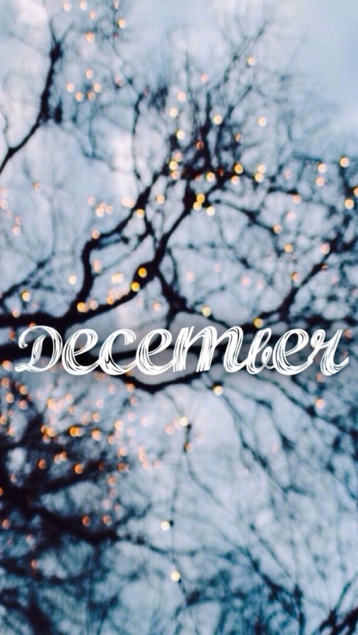 December written in white cursive font, over a blurry background, featuring a tree with white branches, and lights in the background - December