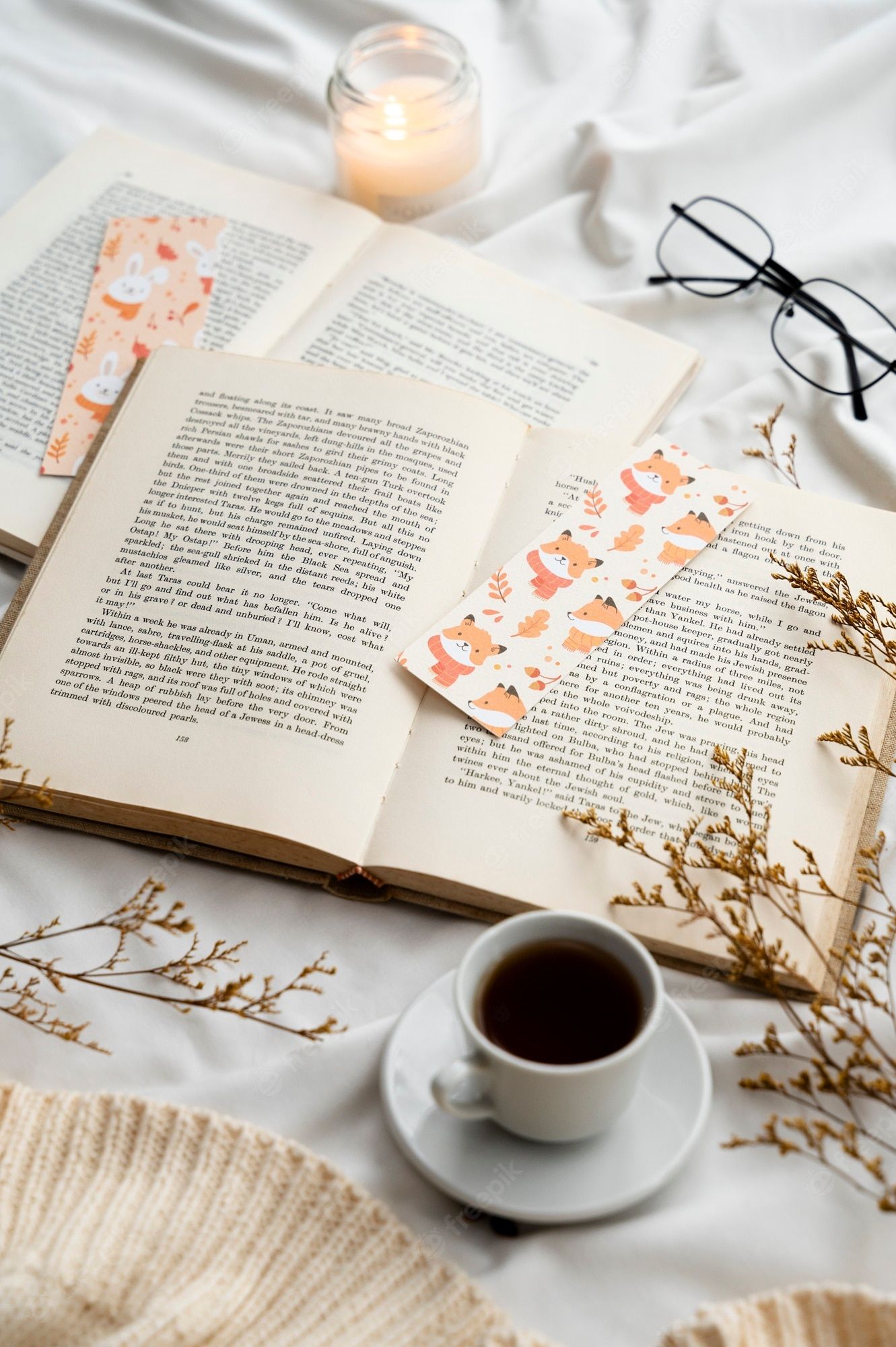 An open book with a fox bookmark, a cup of coffee, and a lit candle on a white table. - Books