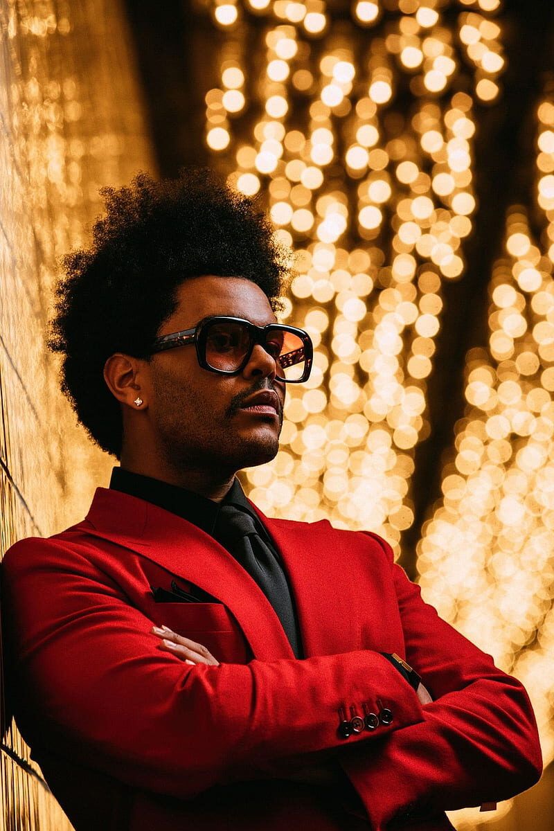 The Weeknd poses in a red suit and sunglasses in front of a wall of lights. - The Weeknd