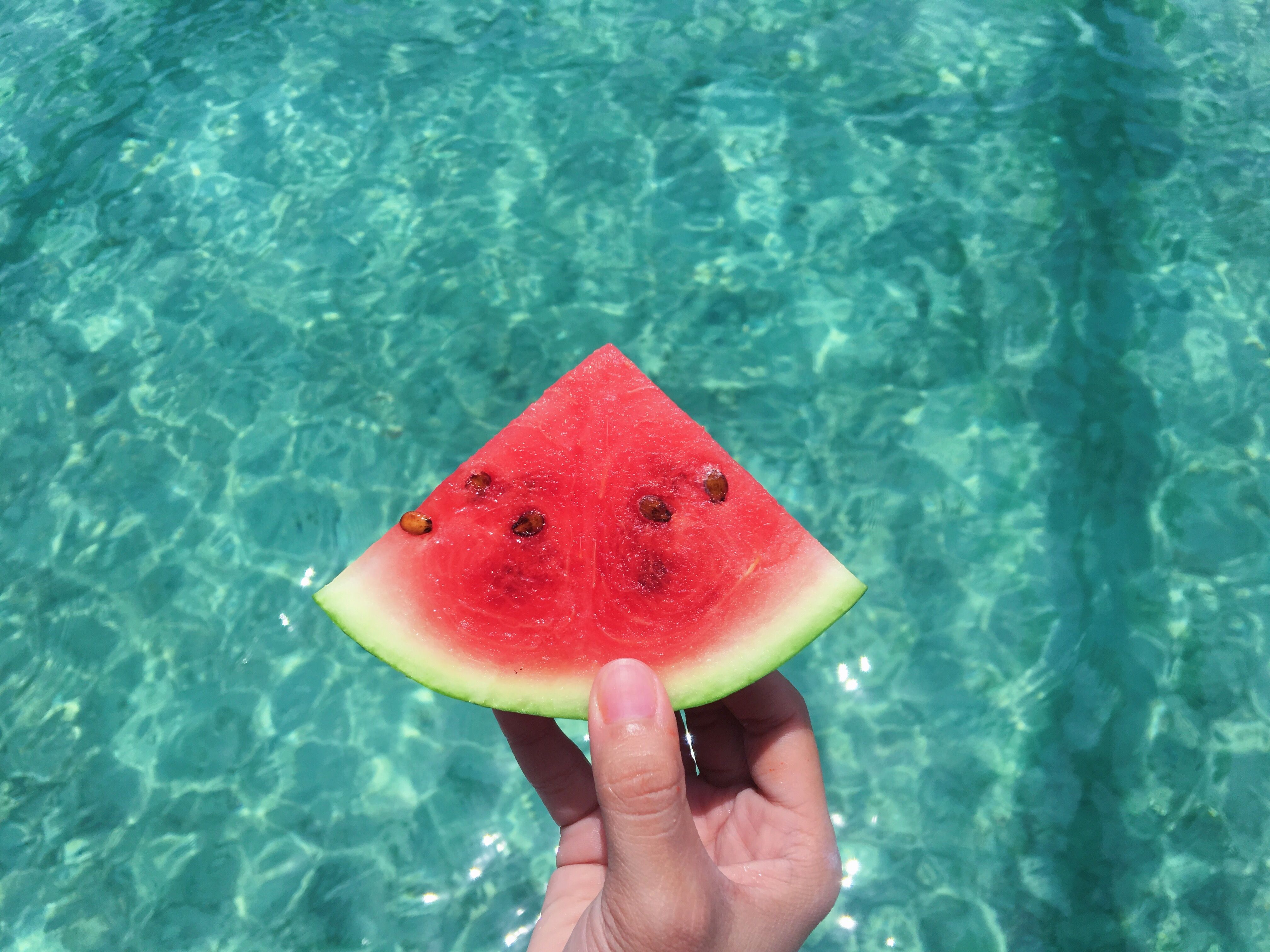 Hand Holding a Slice of Watermelon With Blue Swimming Pool Water in the Background · Free