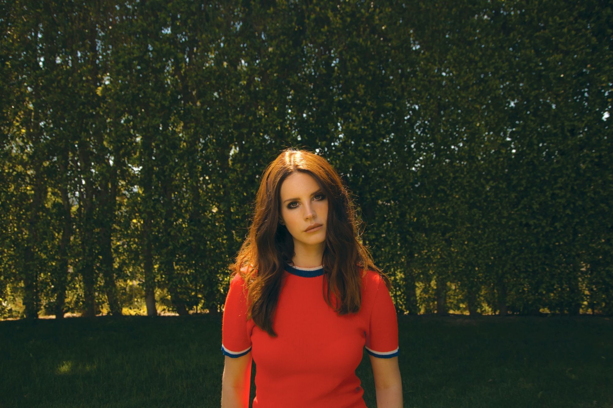 A woman in a red shirt standing in front of a tree. - Lana Del Rey