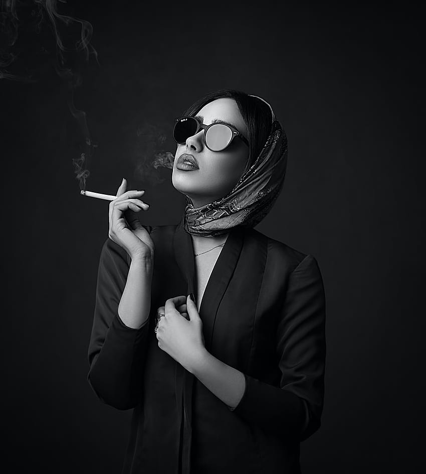 A woman wearing glasses and a headscarf smokes a cigarette. - Smoke