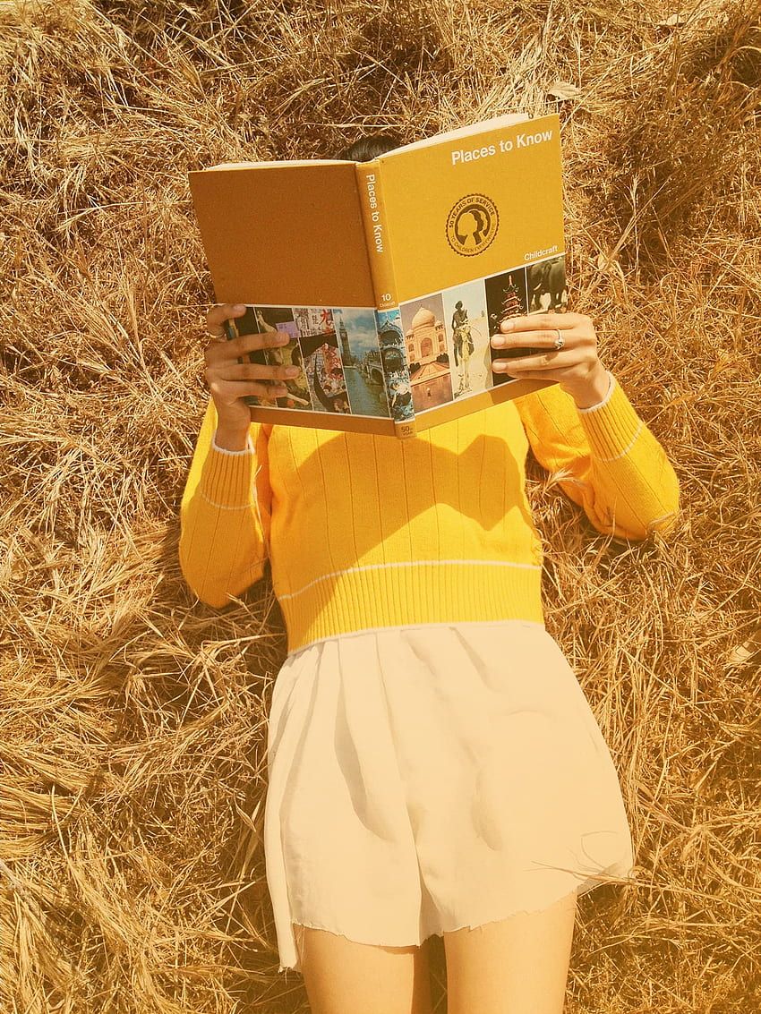 A woman in a yellow top and white shorts lies on a bale of hay, reading a book. - 