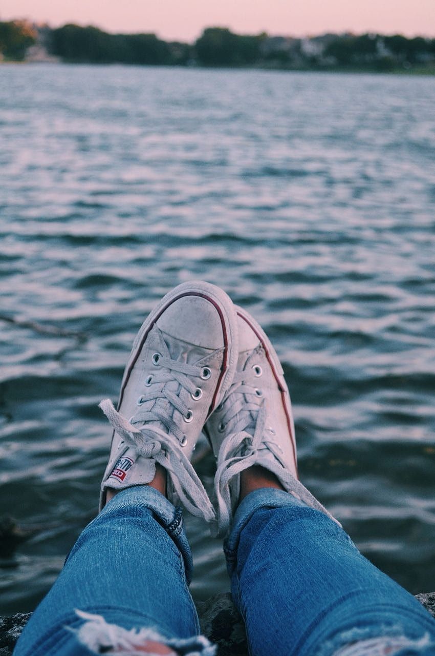 A person wearing converse sneakers and jeans sitting on a rock by the water - Converse