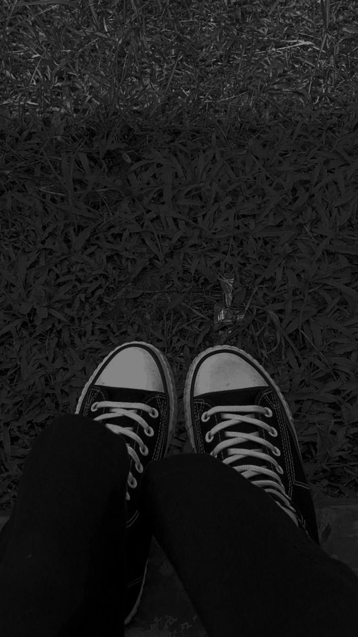 A pair of feet wearing black and white converse sneakers standing on grass. - Converse