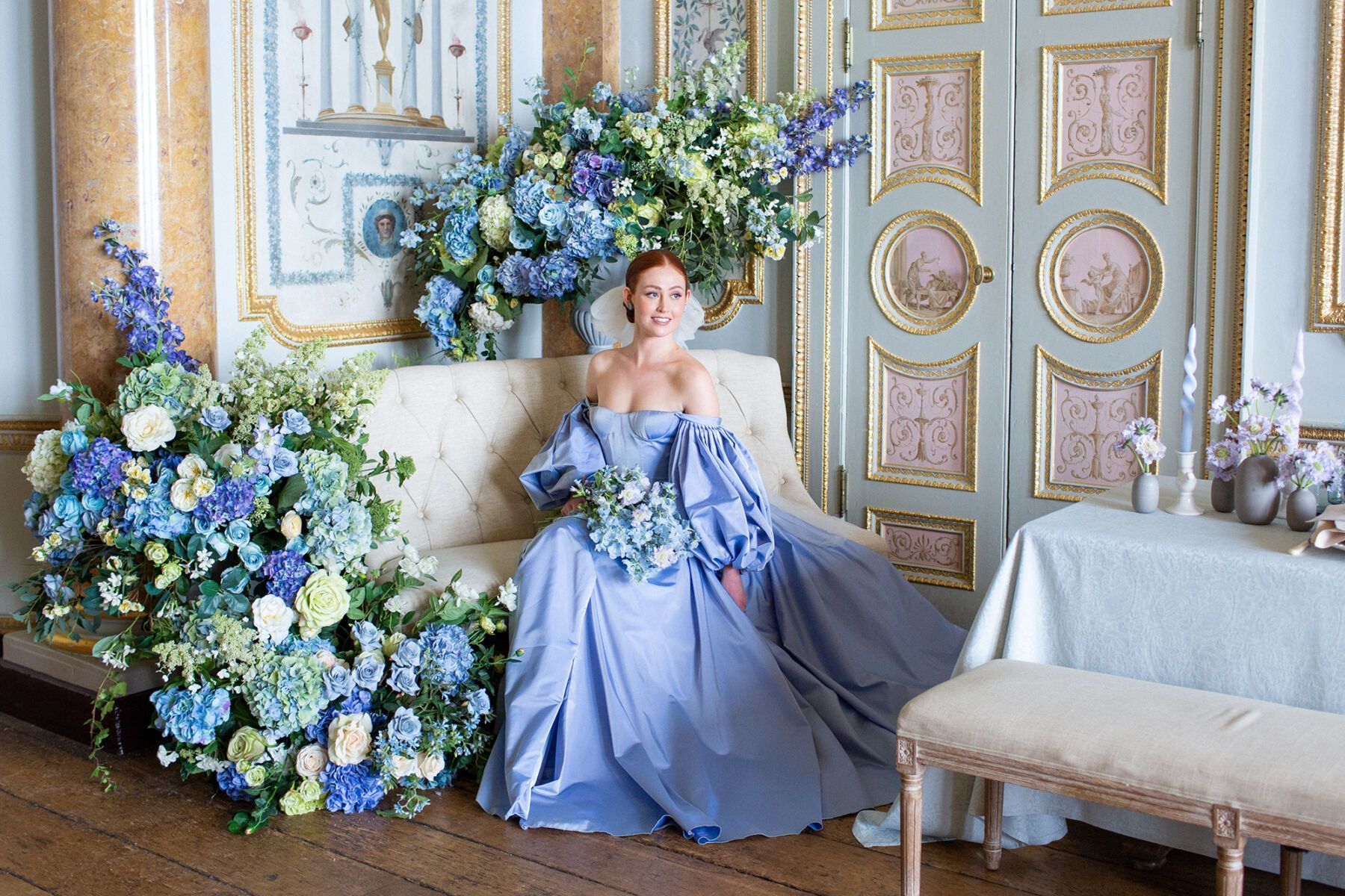 A bride in a blue dress sits on a sofa with a large bouquet of blue flowers. - Bridgerton