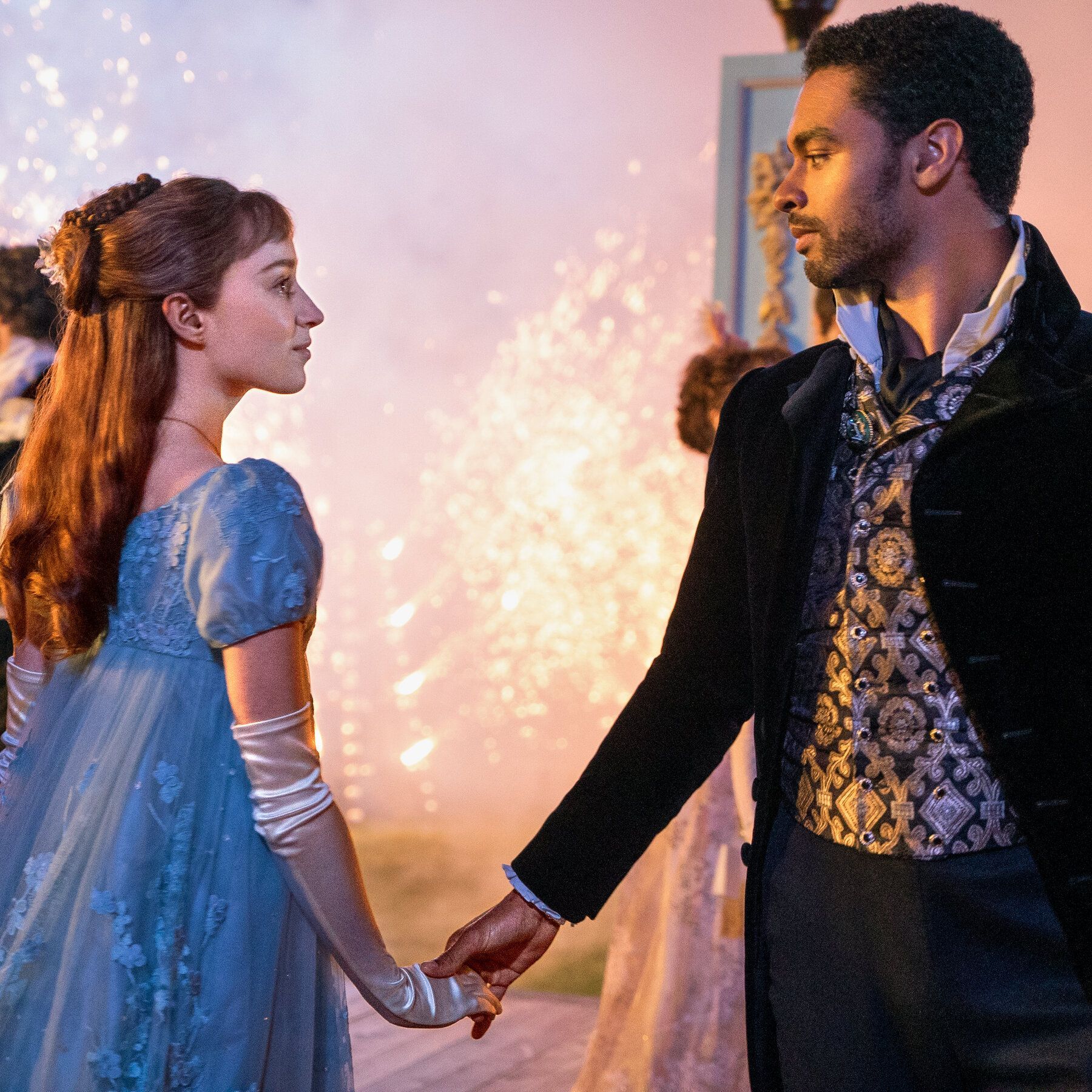 A white woman with red hair and a blue dress holds hands with a black man in a suit in front of a pink background with fireworks - Bridgerton