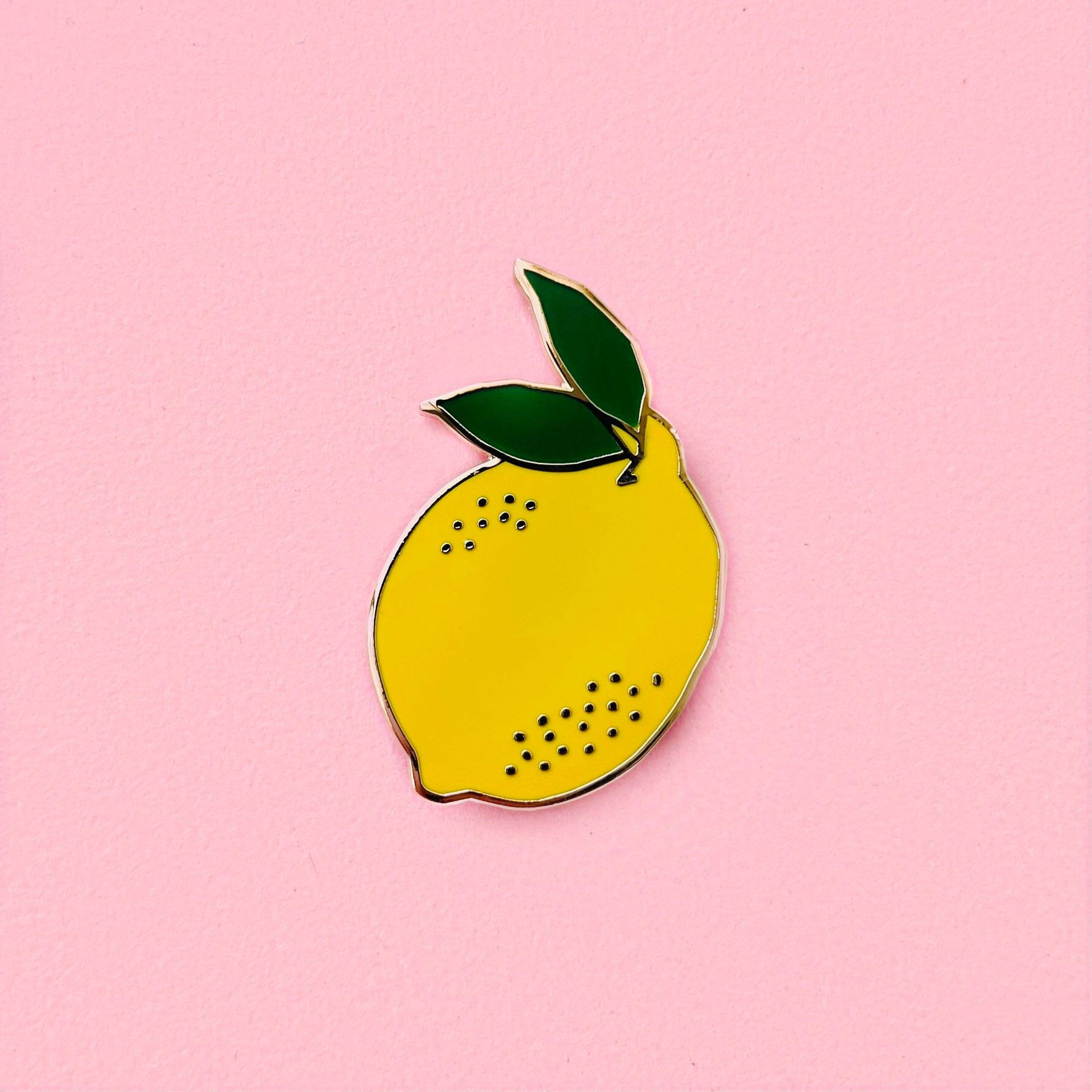 A lemon enamel pin with two leaves on top, all in front of a pink background - Lemon