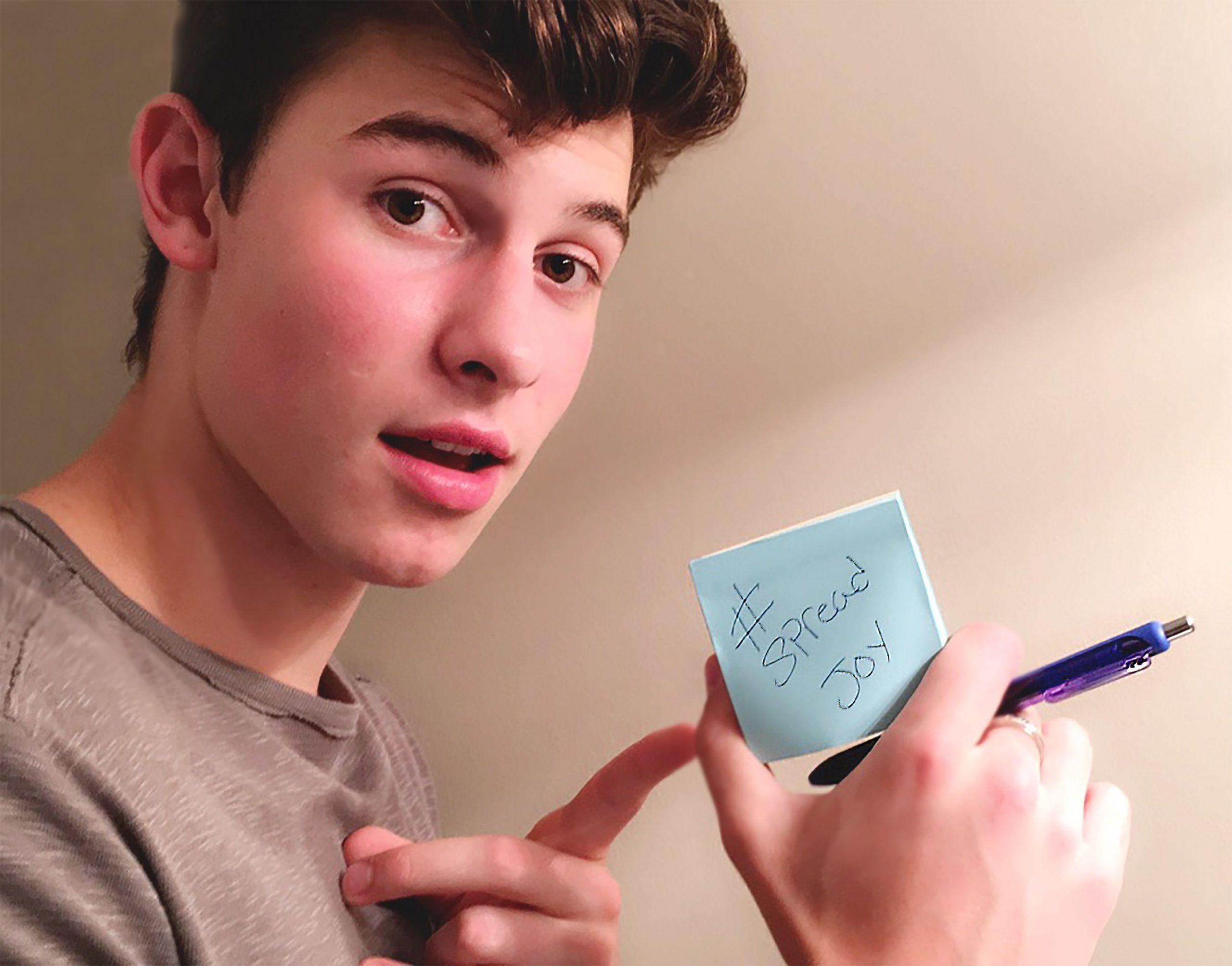A selfie of a young man holding a pen and a post-it note with the hashtag #spreadjoy. - Shawn Mendes