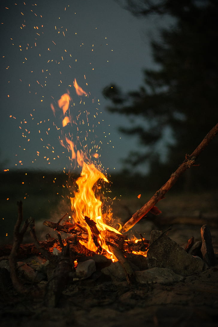 A campfire burns in the dark, with sparks flying out of it. - Camping