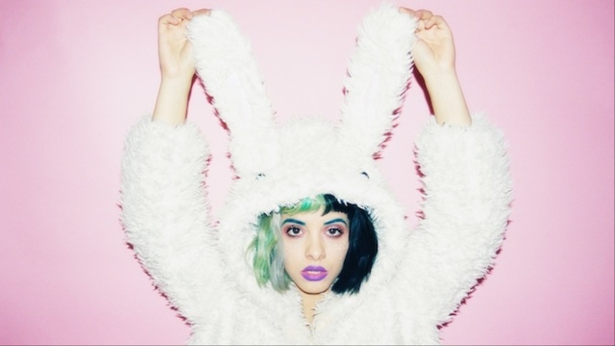 A woman with green and black hair and purple lipstick holds up a white fluffy bunny hat. - Melanie Martinez