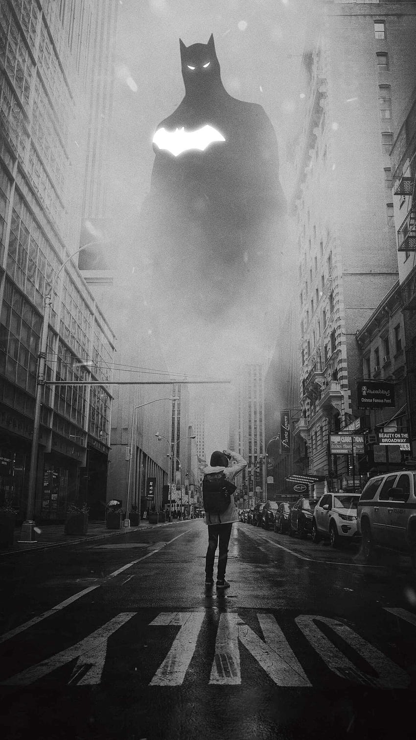 Black and white photo of a man standing in the middle of a city street, with a large batman figure looming above him. - Batman