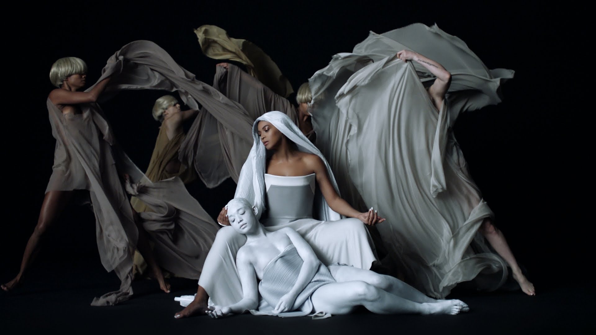 A woman in a white dress sits on the ground with white people in white body casts. They are surrounded by women in white flowing robes. - Beyonce