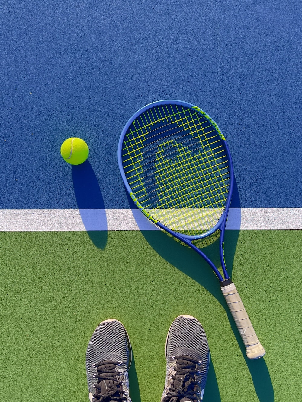 A tennis racket and ball on a tennis court with a person's feet - Tennis