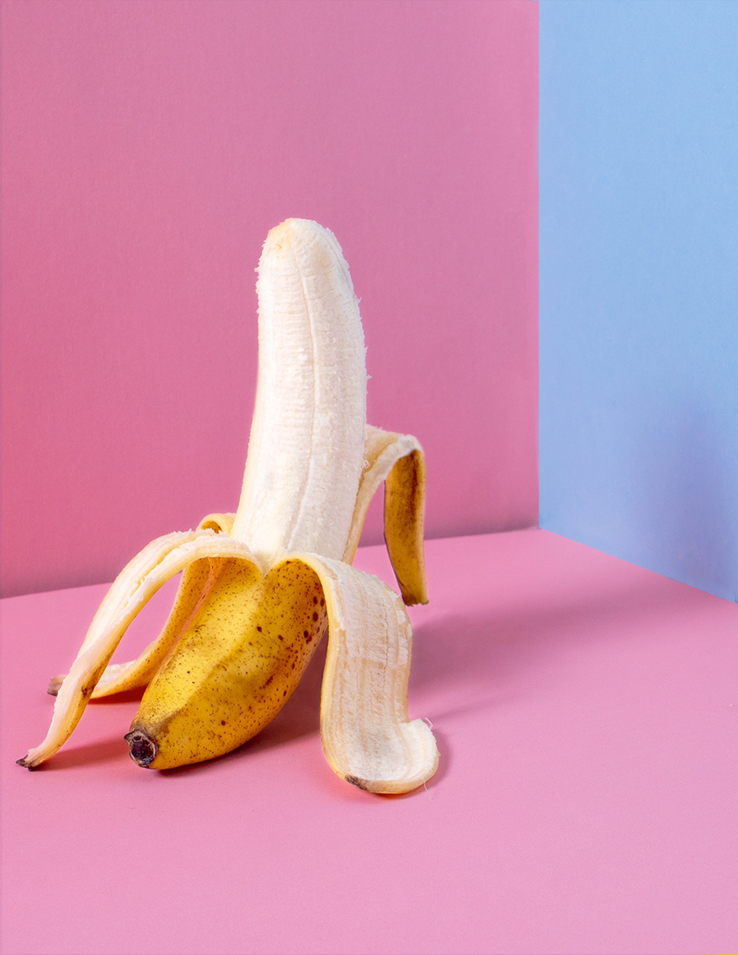 A half peeled banana sits on a pink surface with a blue wall behind it. - Banana