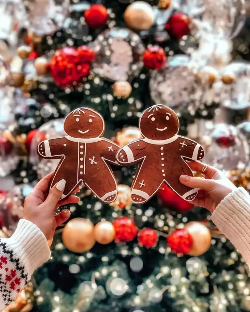 Two gingerbread men held in front of a Christmas tree. - Cozy