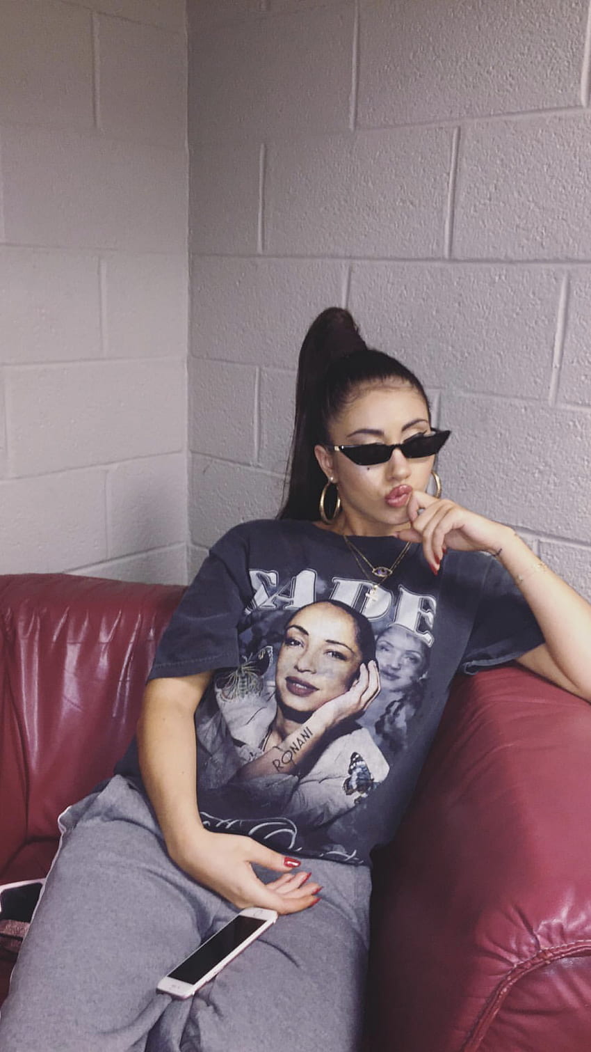 A woman sitting on a couch wearing a Sade t-shirt and sunglasses. - Kali Uchis