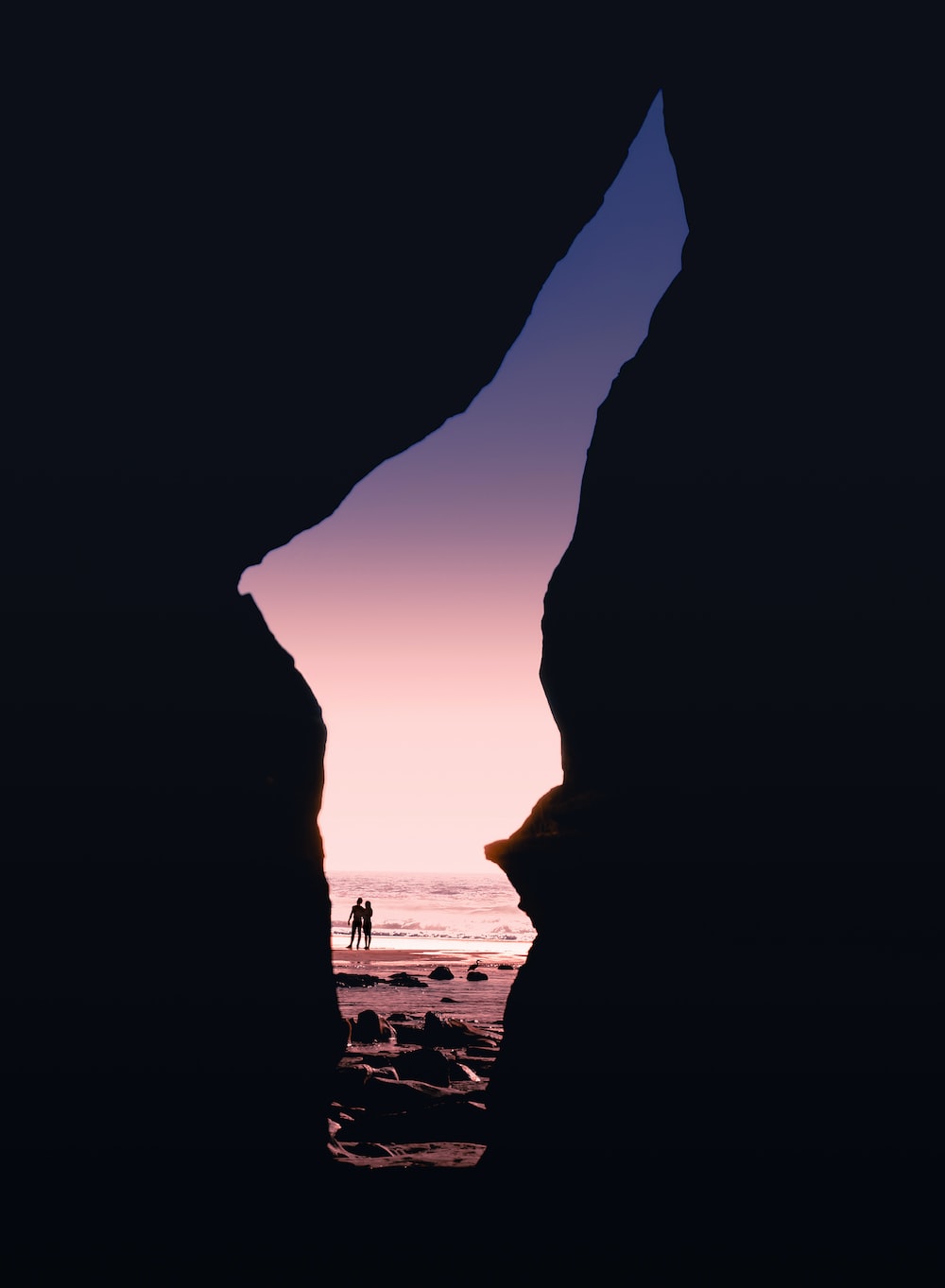A couple standing on a beach at sunset, framed by a large rock formation. - Nature