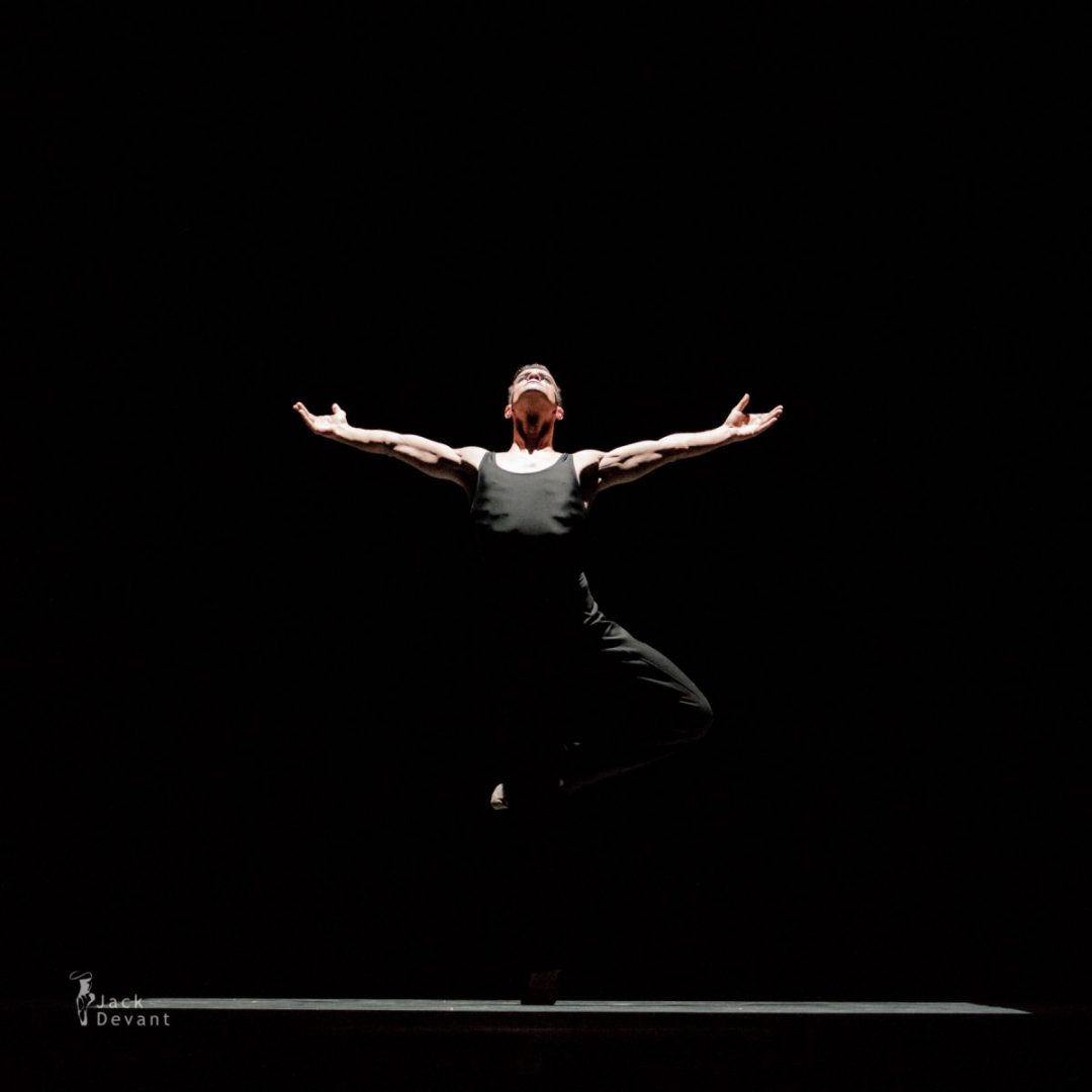Dancer in a black top and black pants, arms outstretched, in a darkened room. - Dance