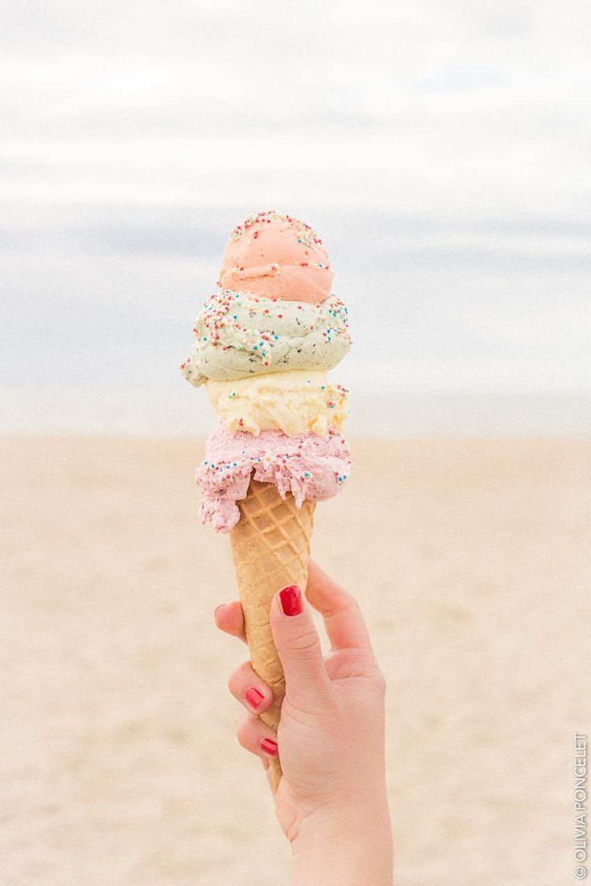 A hand holding a cone with three scoops of ice cream on the beach - Ice cream